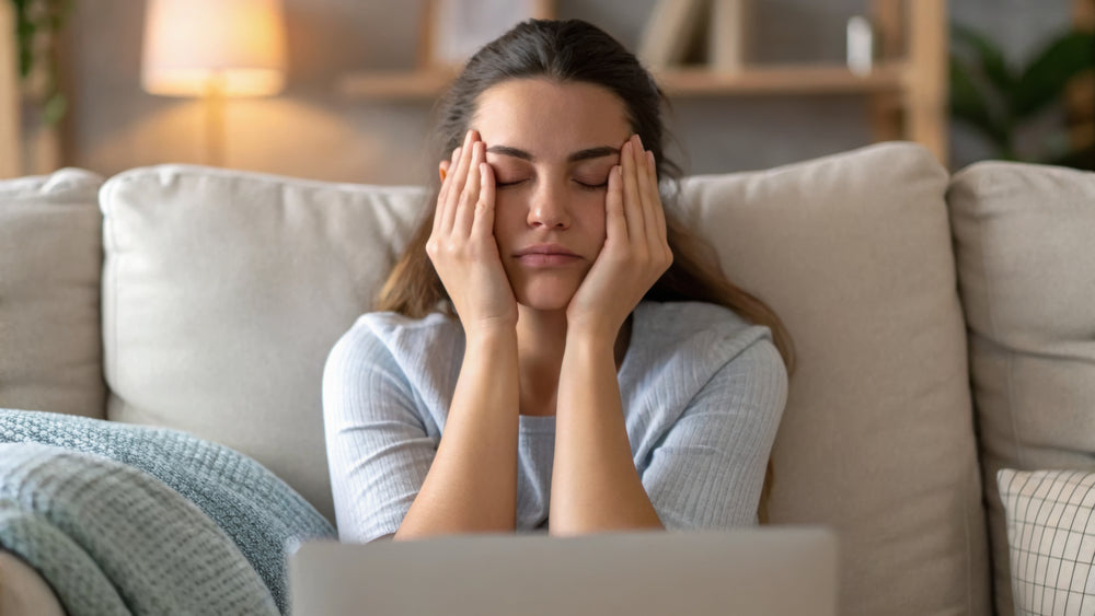 A woman sits on a beige couch with her eyes closed, holding her temples in a gesture of discomfort or stress, with a laptop in the foreground and a softly lit home interior behind her.