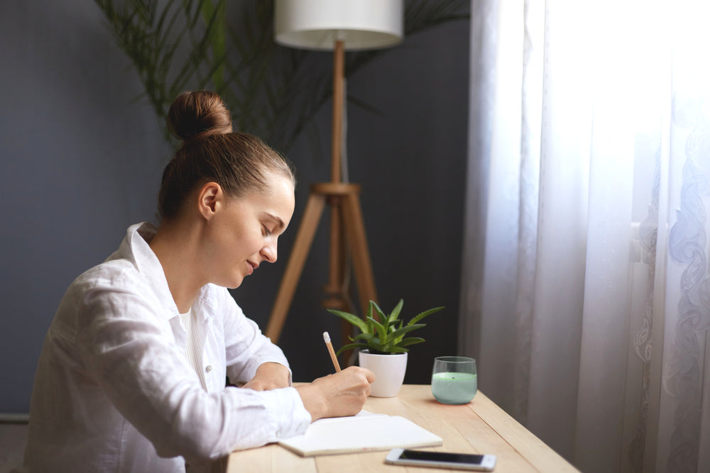 A woman with her hair in a bun sits at a wooden desk, writing in a notebook with a pencil. She is wearing a white button-up shirt.